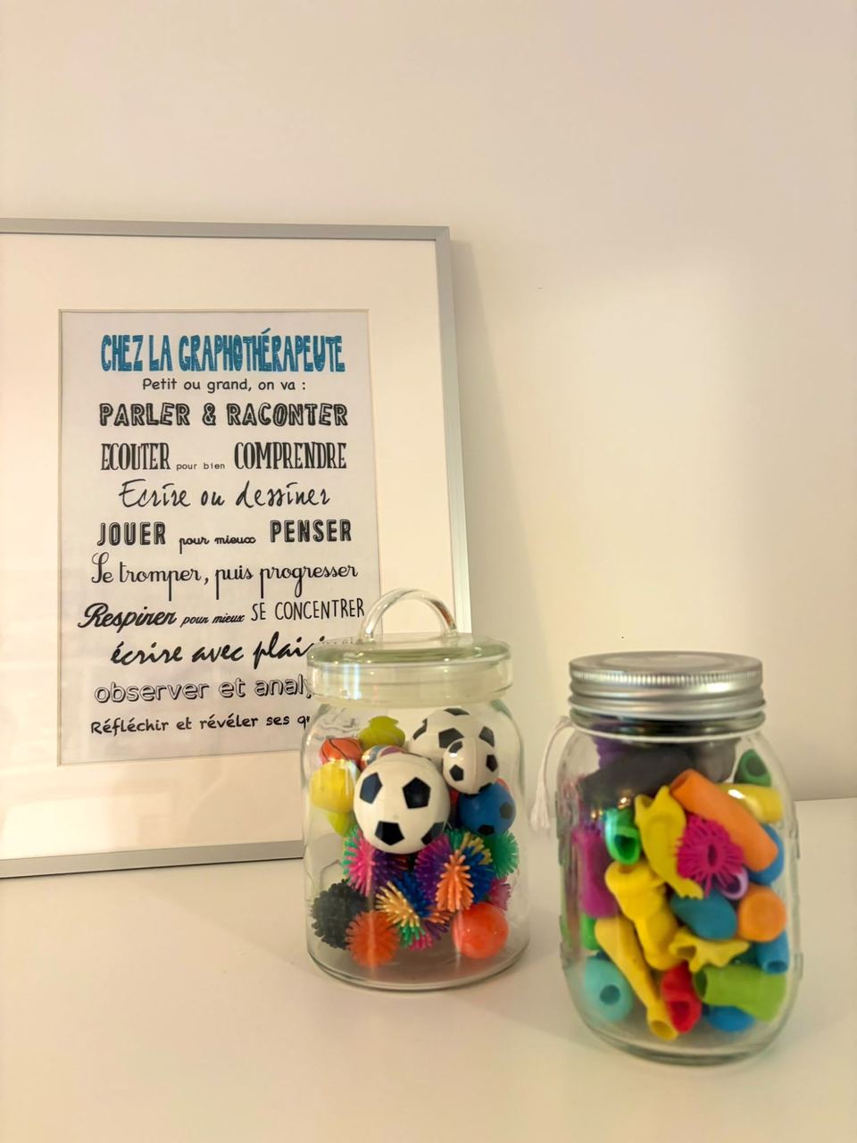 Affiche « Chez la graphothérapeute » et bocaux remplis de balles et de guide-doigts colorés