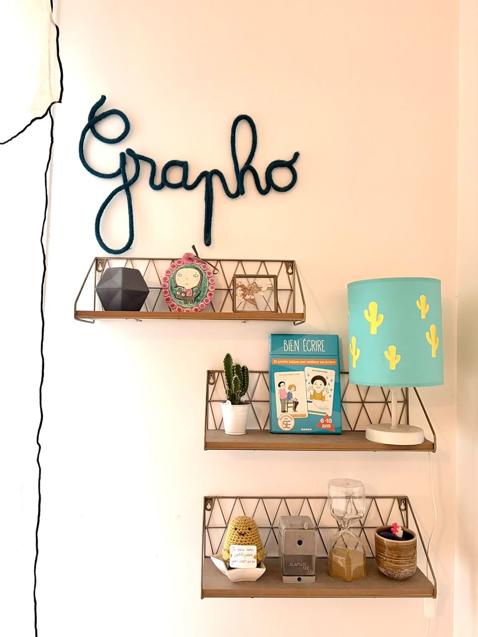 Mur décoré du mot « Grapho » en lettres de laine, étagères avec lampe et livre « Bien écrire »