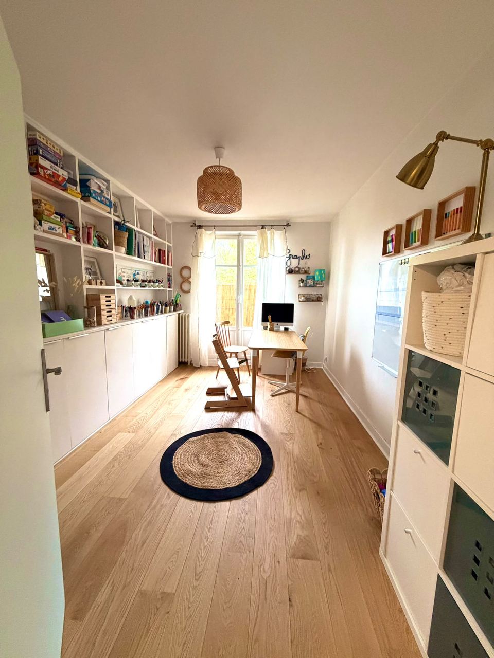 Salle du cabinet : parquet clair, bureau et chaise réglable devant la fenêtre, étagères de jeux