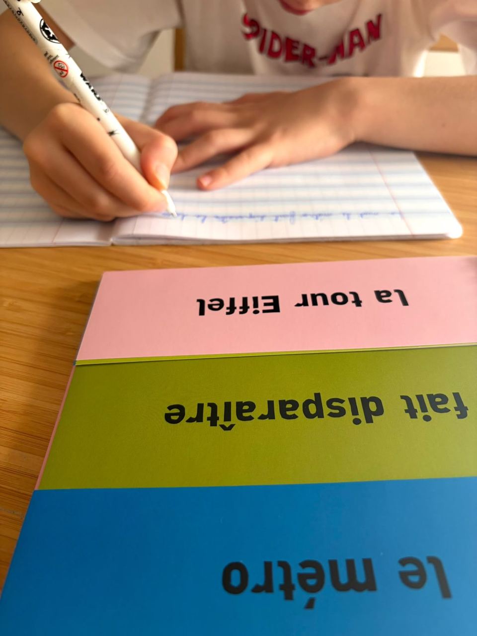 Enfant écrivant dans un cahier, devant des cartes « le métro », « fait disparaître », « la tour Eiffel »