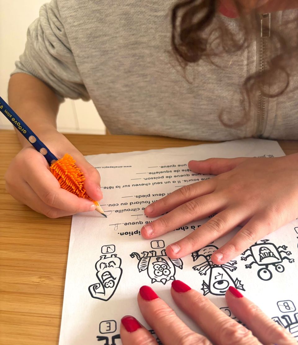 Enfant écrivant avec un crayon muni d'un guide-doigts orange, l'autre main tenant la feuille
